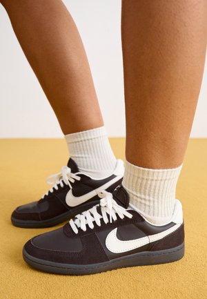 Fekete-fehér Nike tornacipők velúrból és bőrből készült felsőrészével, fehér fűzővel, bordázott krém színű zoknival viselve, sárga háttér előtt.