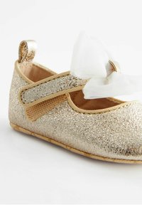 Złote, brokatowe buty Mary Jane z białą tkaninową kokardą i paskiem. Miękki materiał o fakturowanym wykończeniu i płaskiej podeszwie.
