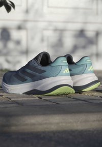 Sportschoenen met een bovenwerk van blauw en grijs mesh, witte tussenzool en groene buitenzool. Drie zwarte strepen aan de zijkanten. Getextureerd en gesculpt design.