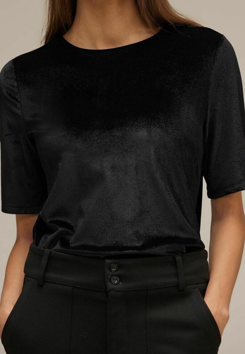 Femme portant un haut en velours noir à manches courtes, rentré dans un pantalon noir taille haute avec fermeture bouton et mains dans les poches.