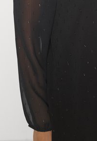 Black blouse with sheer puff sleeves, featuring subtle, reflective dots throughout the fabric. Soft texture with a relaxed fit.
