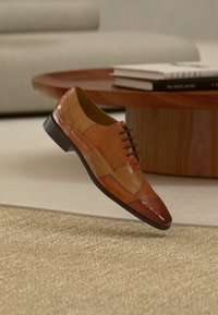 Καφέ δερμάτινο παπούτσι κεντημένο με κομμάτια που αιωρείται πάνω από μπεζ χαλί, με ξύλινο τραπεζάκι καφέ και στοίβες βιβλίων στο φόντο.