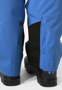 Modré voděodolné kalhoty s černými akcenty a značkou Helly Tech, které mají hladkou texturu a přiléhavý tvar kolem kotníků.