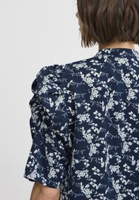 Navy floral blouse with short puff sleeves, featuring white botanical patterns and a smooth fabric texture. Back view shows detailing on shoulders.