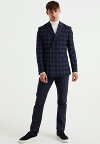 Marineblaues kariertes Doppelbrust-Blazer mit Reverskragen, kombiniert mit marineblauen Hosen und schwarzen Wildledersneakern, darunter ein cremefarbener Rollkragenpullover.