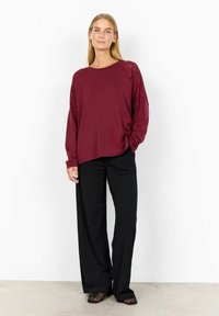Woman standing wearing a burgundy long-sleeve top with lace shoulders, black wide-leg pants, and black mesh shoes against a white background.