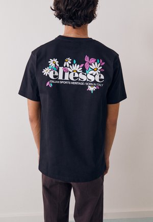 Person, die ein schwarzes T-Shirt mit weißem "ellesse"-Schriftzug trägt, umgeben von weißen Gänseblümchen und rosa Blättern, steht vor einer schlichten hellen Wand.