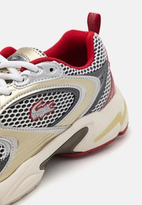 Sneaker featuring a mesh upper in white, grey, and red with leather accents. Lace closure and cushioned sole, highlighted by a logo detail.