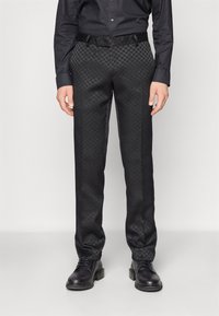 Černé kalhoty se vzorem a texturovaným povrchem, s přizpůsobeným střihem a tkaným černým opaskem. Doplňují je černé kotníčkové boty.