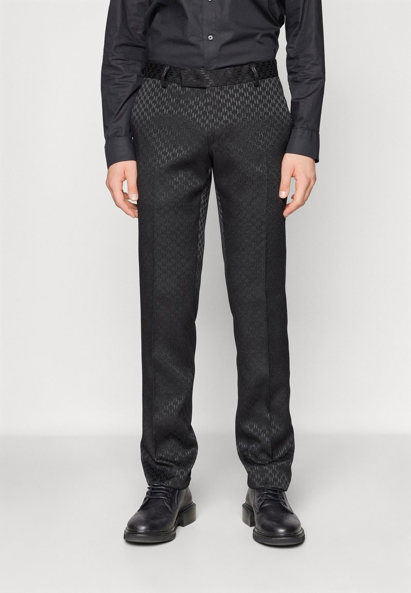 Černé kalhoty se vzorem a texturovaným povrchem, s přizpůsobeným střihem a tkaným černým opaskem. Doplňují je černé kotníčkové boty.