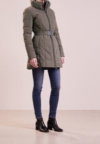 Olivgrön vadderad jacka med midjebälte, hög krage och dragkedja fram; tillsammans med blå skinny jeans och svarta ankelstövlar.