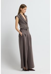 Nátělník bez rukávů s hlubokým výstřihem do V v hnědém texturovaném materiálu s páskem v pase a kalhotami s širokými nohavicemi. Má zapínání na jeden knoflík.