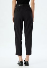 Hoge taille zwarte broek met een getailleerde pasvorm, voorzien van plooien, een riem en een achterzak. Gecombineerd met zwarte gestructureerde schoenen.