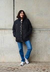 Čierna prešívaná bunda s vysokým golierom a zipsovými vreckami, skombinovaná s pruhovanou košeľou, modrými džínsy a bielymi teniskami pri betónovej stene.