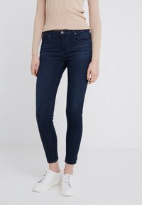 Mörkblå smala jeans med framlommer och fem bälteshål, i kombination med en ljuskampad beige tröja och vita sneakers.