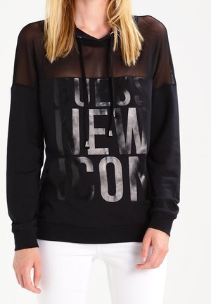 Kvinde iført en sort hættetrøje med gennemsigtigt øverste panel og skinnende tekst, kombineret med hvide bukser, stående mod en ensfarvet baggrund.