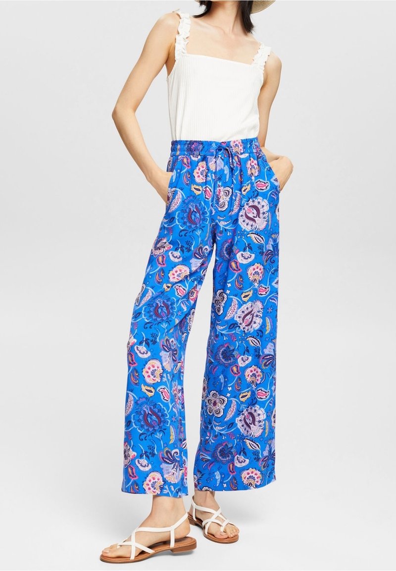 Femme portant un haut sans manches blanc à volants, un pantalon large bleu avec un motif floral coloré et des sandales blanches à brides.