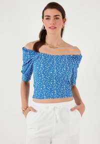 Blå off-shoulder top med små hvide blomster mønstre, korte puffede ærmer og en tætsiddende pasform, parret med hvide bukser med elastisk talje.