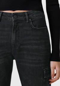 Jeans de mezclilla negra con una textura suave, cintura alta, cierre de botón y bolsillo lateral tipo cargo. Un sutil detalle de costura realza el diseño.