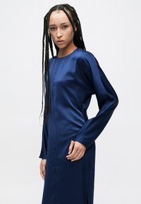 Tengerészkék szatén ruha hosszú ujjakkal, kerek nyakkivágással, testhez simuló szabással és oldalsó sliccel; sima textúrájú, minimalista dizájn.