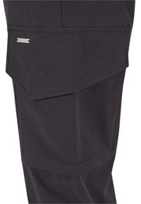 Fekete szövetes cargo zseb döntött fedéllel és kis téglalap alakú fém logócímkével a nadrág lábán.