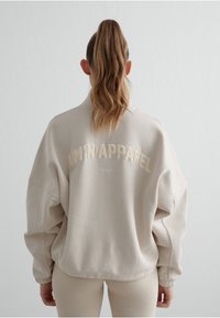 Béžová oversized mikina s vysokým límcem, na zadní straně s vyšitým textem "AIM'N'APPAREL Sport". Měkká textura a volný střih.