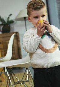Jasny, dzianinowy sweter z okrągłym wzorem w kolorze pomarańczowym i zielonym. Dziecko trzyma żółte jabłko, ubrane w ciemne spodnie. Krzesło w tle.