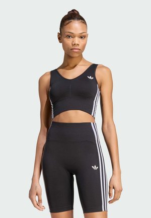 Vrouw die een zwarte Adidas mouwloze crop top en bijpassende hoge taille biker shorts met witte strepen aan de zijkanten draagt, staand tegen een effen achtergrond.