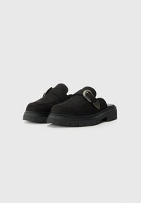 Črne semiš slip-on čevlje z debelim, teksturiranim podplatom in kovinskim detajlom na traku. Okrogla konica in minimalistična zasnova.