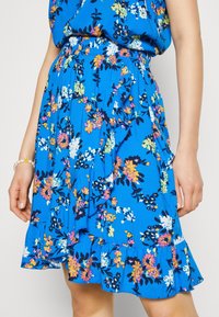 Sininen kukkaroikkuva hame kevyestä kankaasta, jossa on kirkkaanvärisiä kukkia vaaleanpunaisena, keltaisena ja valkoisena, rypytetyllä helmapäällä ja joustavalla vyötäröllä.