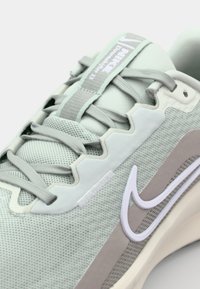 Lys grøn atletiksko har en mesh-overdel, teksturerede snørebånd og et hvidt Nike swoosh-logo langs siden. Gummisål med beige accenter.