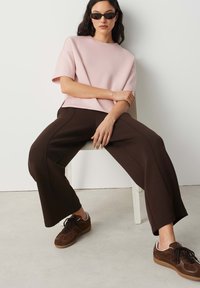 Frau sitzt auf einem Stuhl, trägt ein hellrosa kurzärmeliges Oberteil, weite dunkelbraune Hose, braune Sneakers und schwarze Sonnenbrille vor einer weißen Wand.
