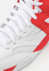 White athletic sneaker featuring red accents, leather and mesh materials, perforated details, and white laces with a padded ankle support.