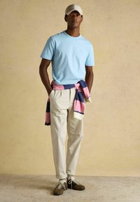 Homme portant un t-shirt bleu clair, un pantalon beige, un pull rayé rose et bleu marine noué autour de la taille, une casquette beige et des baskets marron, debout devant un fond uni.