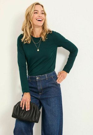 Grünes Langarmoberteil, kombiniert mit hochgeschnittenen, weit geschnittenen blauen Jeans. Hält eine schwarze gefaltete Clutch. Einfache Halskette als Akzent.