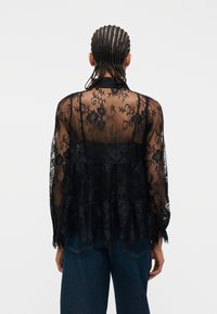 Άτομο με πλεκτά μαλλιά που φοράει διαφανές μαύρο δαντελένιο μπλουζάκι και σκούρο μπλε τζιν, εμφανιζόμενο από την πίσω πλευρά πάνω σε απλό λευκό φόντο.