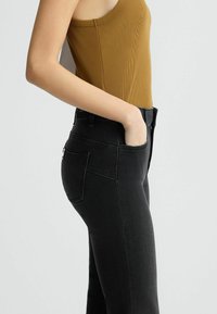 Jean noir taille haute à coupe ajustée, avec des poches arrière et un accent en métal doré à la taille. Associé à un débardeur marron côtelé.