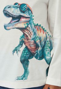 Weißes Langarmhemd mit einem bunten, detaillierten Grafik eines T-Rex, der orangefarbene Sonnenbrille trägt, mit sichtbaren Stacheln und Klauen.