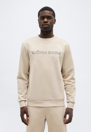 Muž ve světle béžové mikině Björn Borg a odpovídajících kalhotách stojí čelem k divákovi na bílém pozadí.