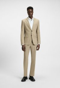 Costume beige ajusté avec un bouton unique, revers crantés et deux poches avant, associé à une chemise blanche et des chaussures noires. Texture lisse.