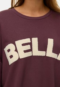 Burgundia huppar, millel on suur, tekstureeritud beež "BELLA" trükk. Kujundus sisaldab ümmargust kaelust ja pehmet kangast.