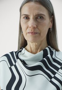 Black and white patterned blouse with smooth fabric, featuring flowing lines and a draped collar, worn with contrasting earrings.