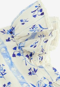 Tissu à motifs floraux dans des nuances de bleu sur un fond crème. Présente des manches volantes et une finition en dentelle. Matériau léger et texturé.