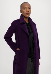 Abrigo morado con solapa con muesca, bolsillos laterales y mangas largas, usado sobre una blusa negra. Textura suave y corte entallado.