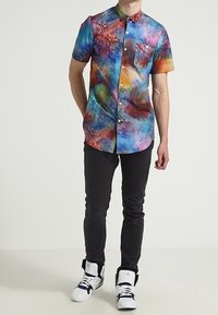 Košile s krátkým rukávem na knoflíky s vícebarevným batikovým vzorem v modré, růžové, červené a zelené barvě. Obsahuje kapsu na hrudi a límec.