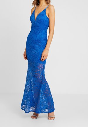 Femme portant une robe longue en dentelle bleu royal avec des bretelles spaghetti et un profond décolleté en V, associée à des talons ouverts argentés.
