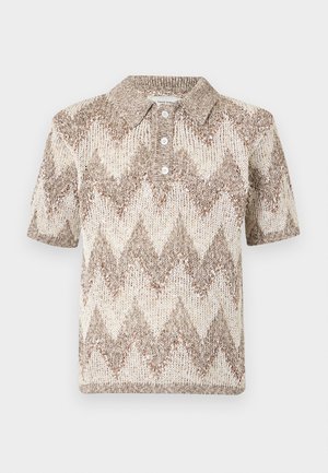 Homme debout portant un polo en maille beige et marron à manches courtes avec motif zigzag, un pantalon marron large et des chaussures noires sur fond blanc.