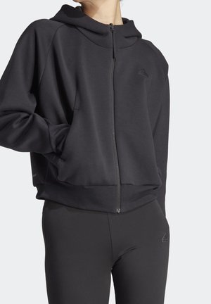Osoba ubrana w czarną bluzę z kapturem na zamek i czarne legginsy z logo Adidas, stojąca z jedną ręką w kieszeni bluzy na jednolitym tle.