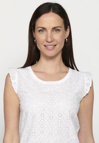 Bílá vyšívaná blůza s květinovými výřezy, krátkými volánkovými rukávy a kulatým výstřihem. Lehcé, strukturované tkaniny s jemnými vzory.