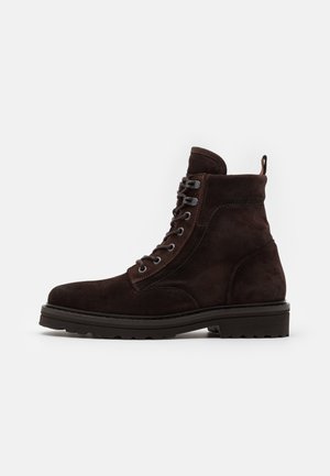 Botte à lacets en daim brun foncé, avec semelle épaisse à crampons et languette à l'arrière, présentée de profil sur fond blanc.